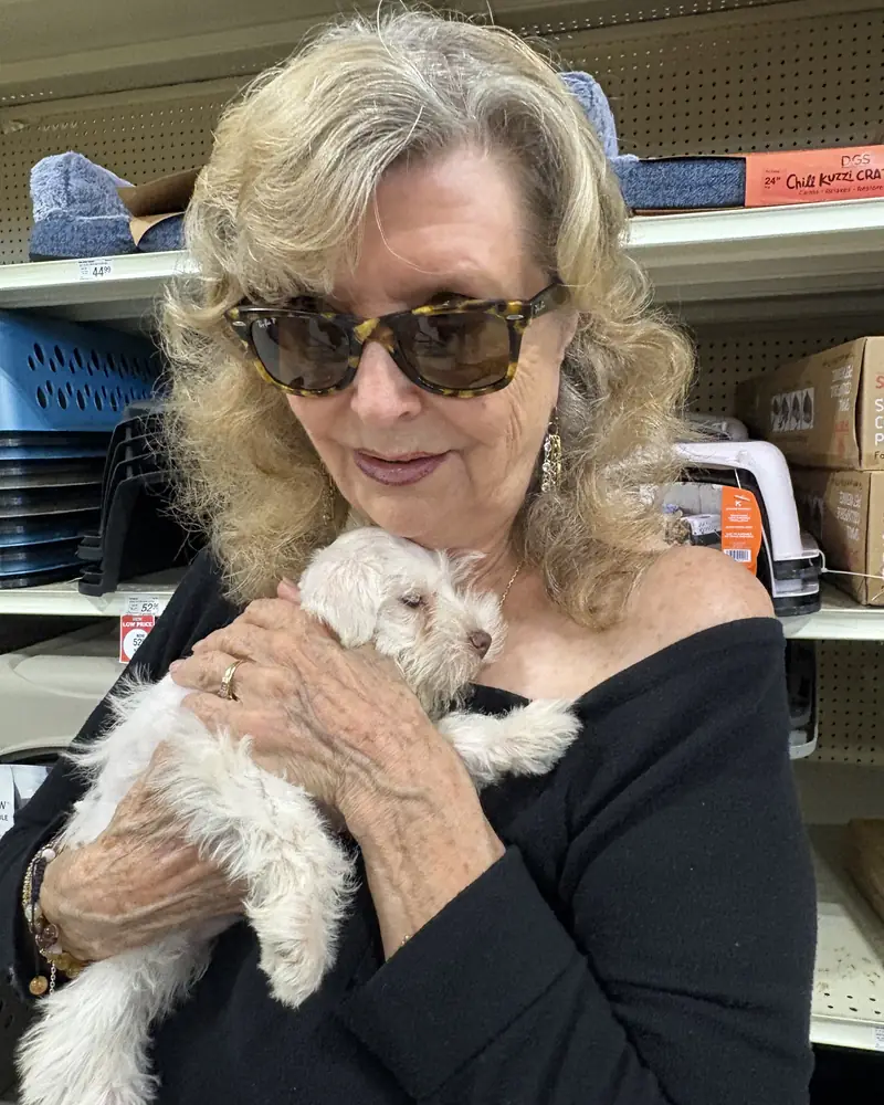 A woman in sunglasses cradling her new white puppy against her shoulder in a pet-supply aisle.