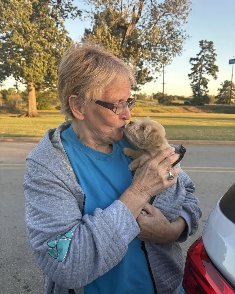 A woman in a grey hoodie leaning in to her new cream puppy, wrapped in the front of her jacket, beside her car.