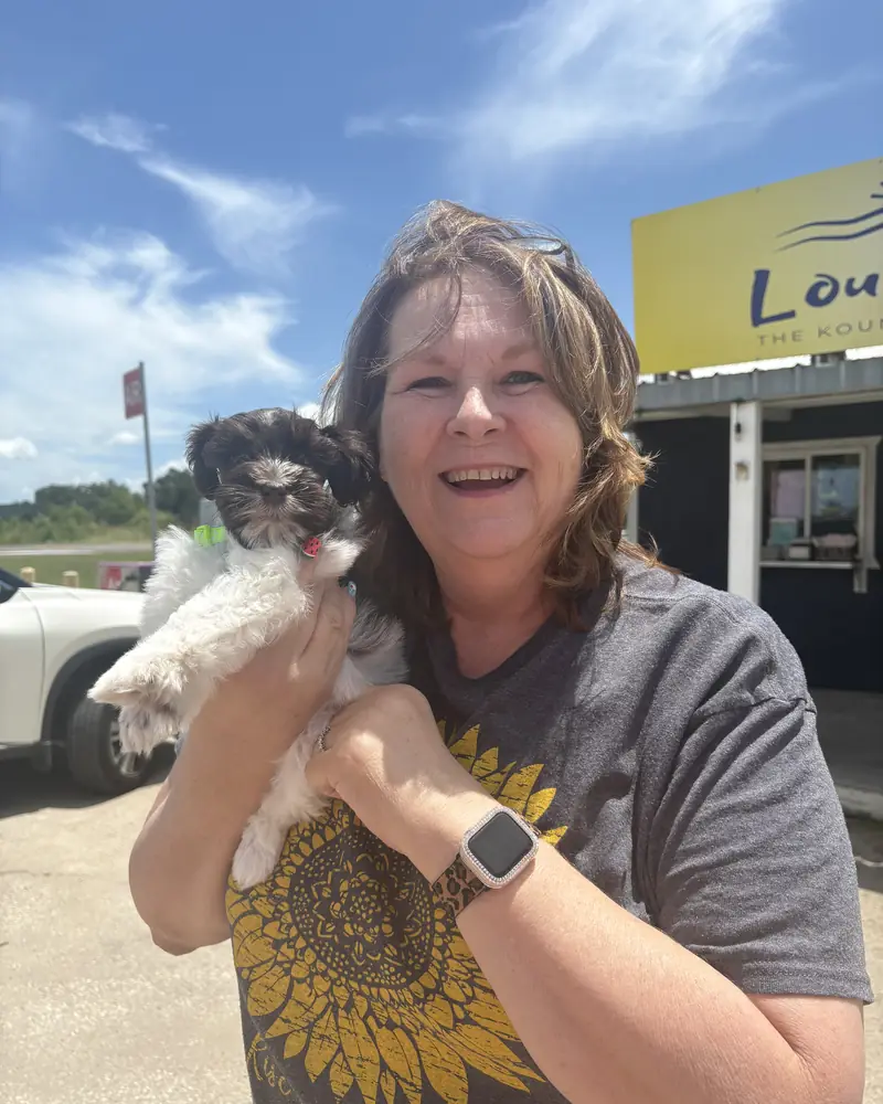 A woman holding up her new black-and-white puppy in both hands outside a roadside shop.