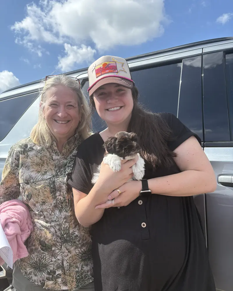 Two women beside a silver van, one holding a new chocolate-and-white puppy, the other carrying a pink blanket.