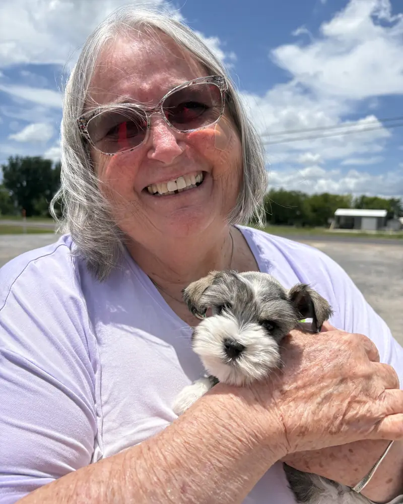 A woman in sunglasses holding her new salt-and-pepper puppy up beside her under a bright summer sky.