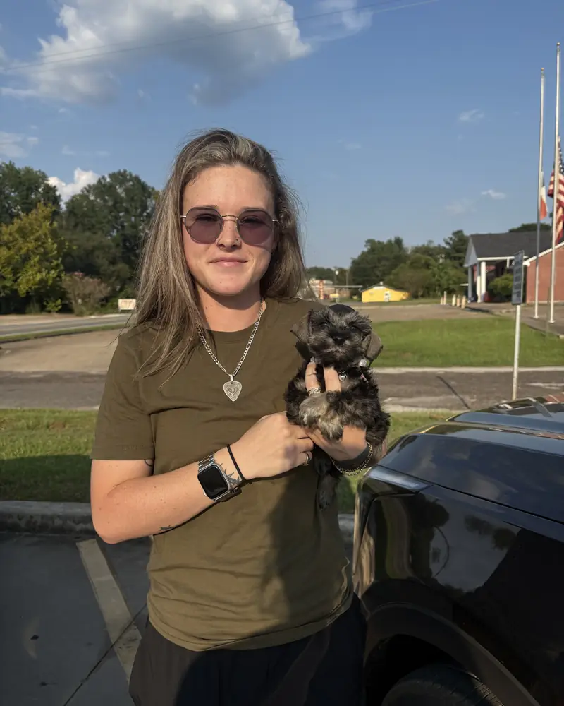 A young woman in an olive T-shirt holding her new black puppy in one hand beside a dark car.