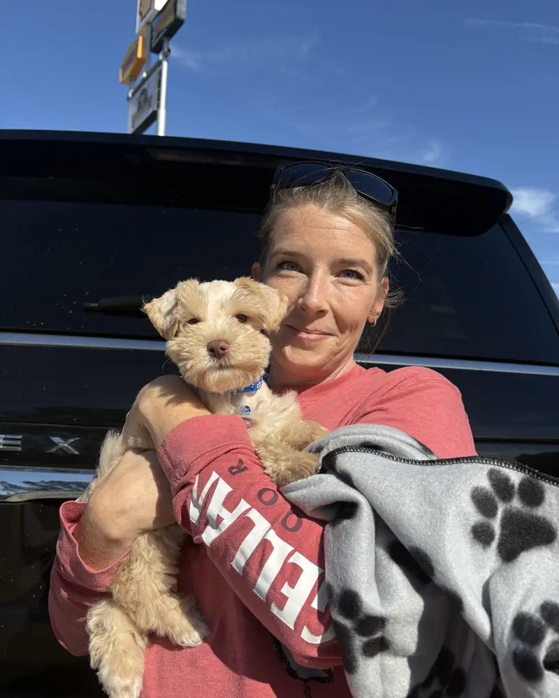 A woman at the back of a black SUV holding her new cream puppy, a paw-print blanket over her arm.