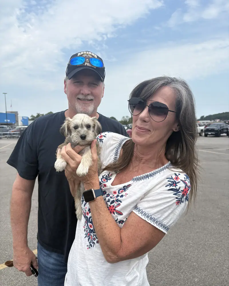 A couple in a car park, the woman holding their new cream puppy up towards the camera.