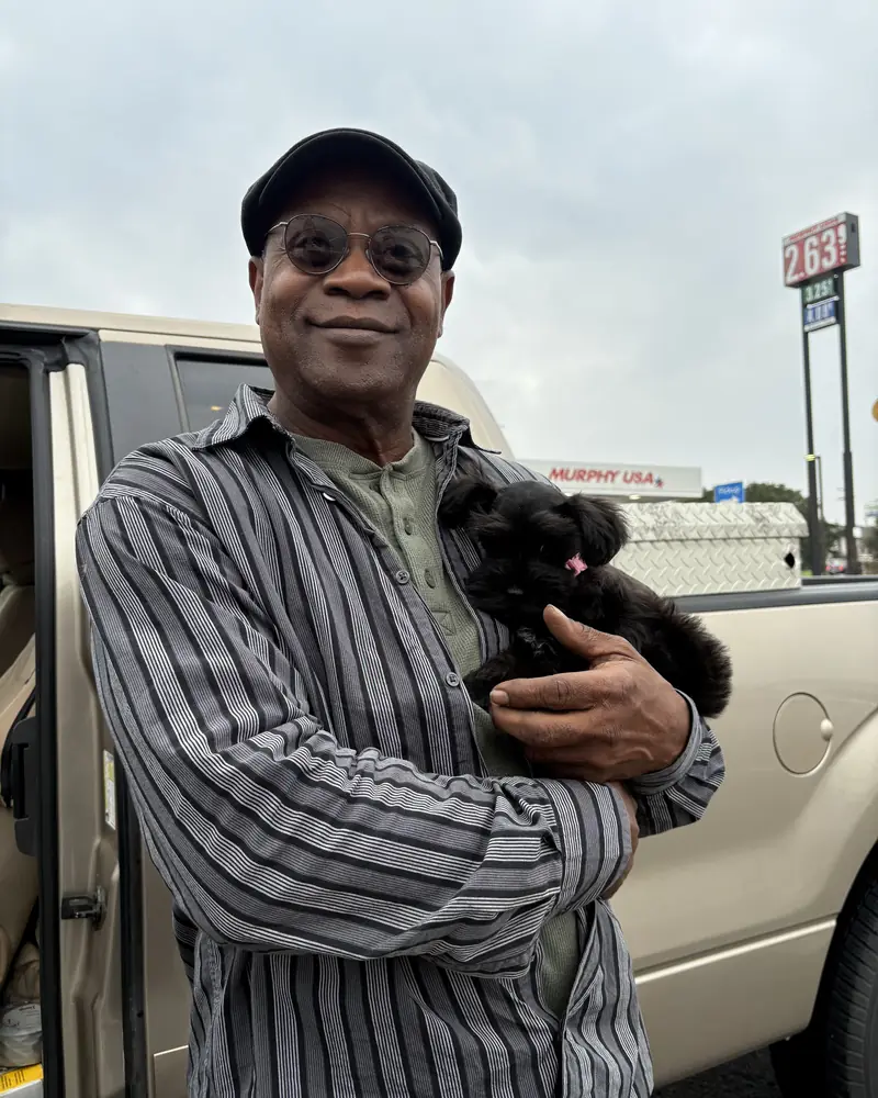 A man in a flat cap holding his new black puppy in the crook of his arm beside his van.