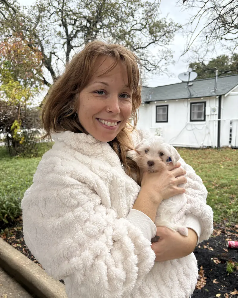 A woman in a cream fleece holding her new white puppy in a garden on a grey autumn day.