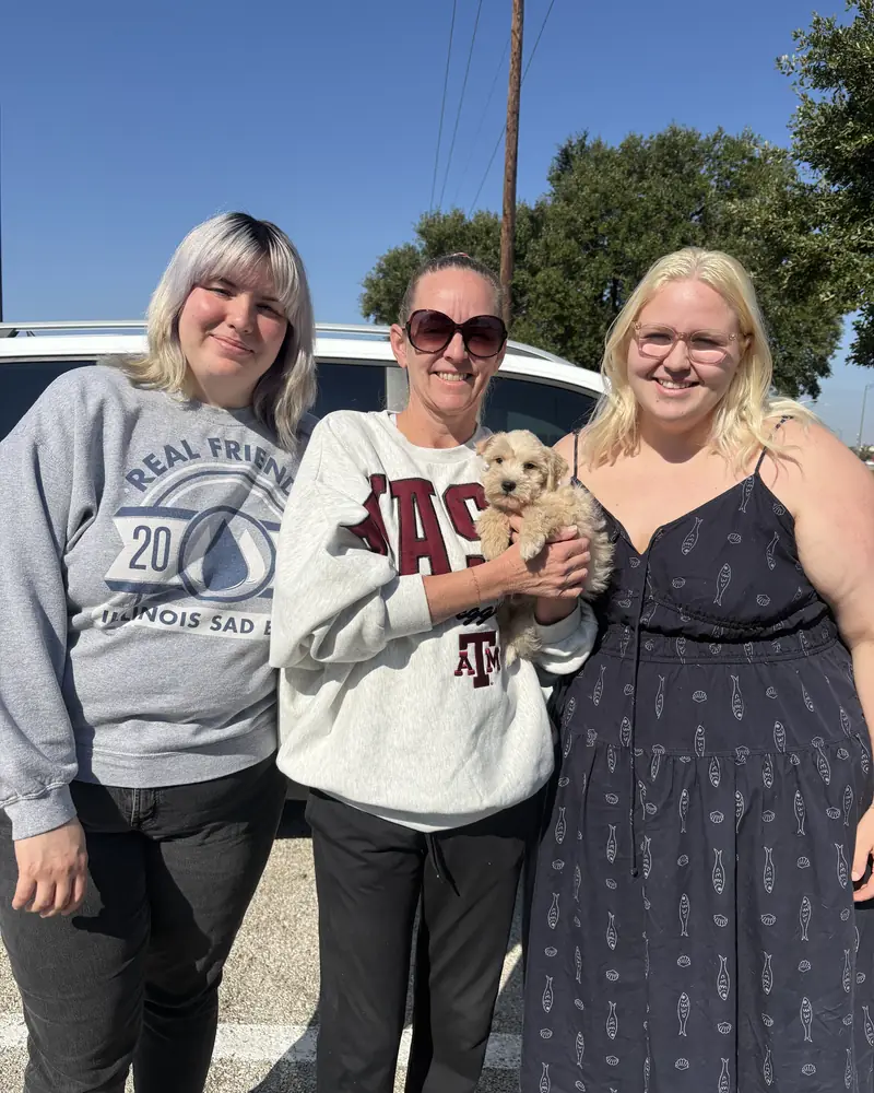 Three women standing side by side in a car park, the one in the middle holding their new cream puppy.