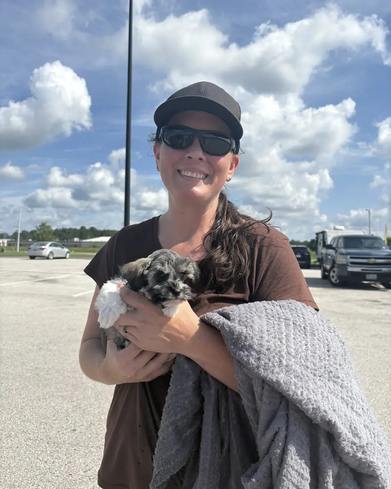 A woman in a cap and sunglasses holding her new salt-and-pepper puppy, a grey blanket over her arm.