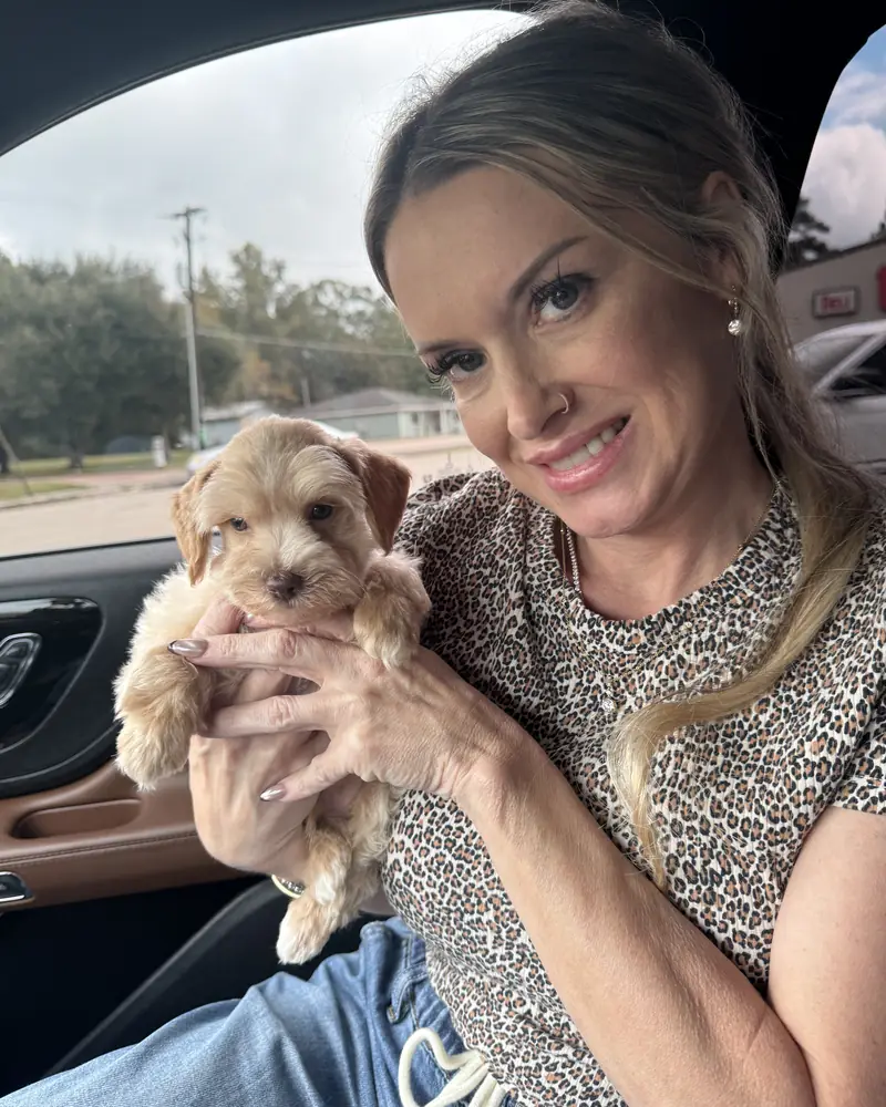 A woman in the passenger seat of a car holding her new cream puppy up to the camera.