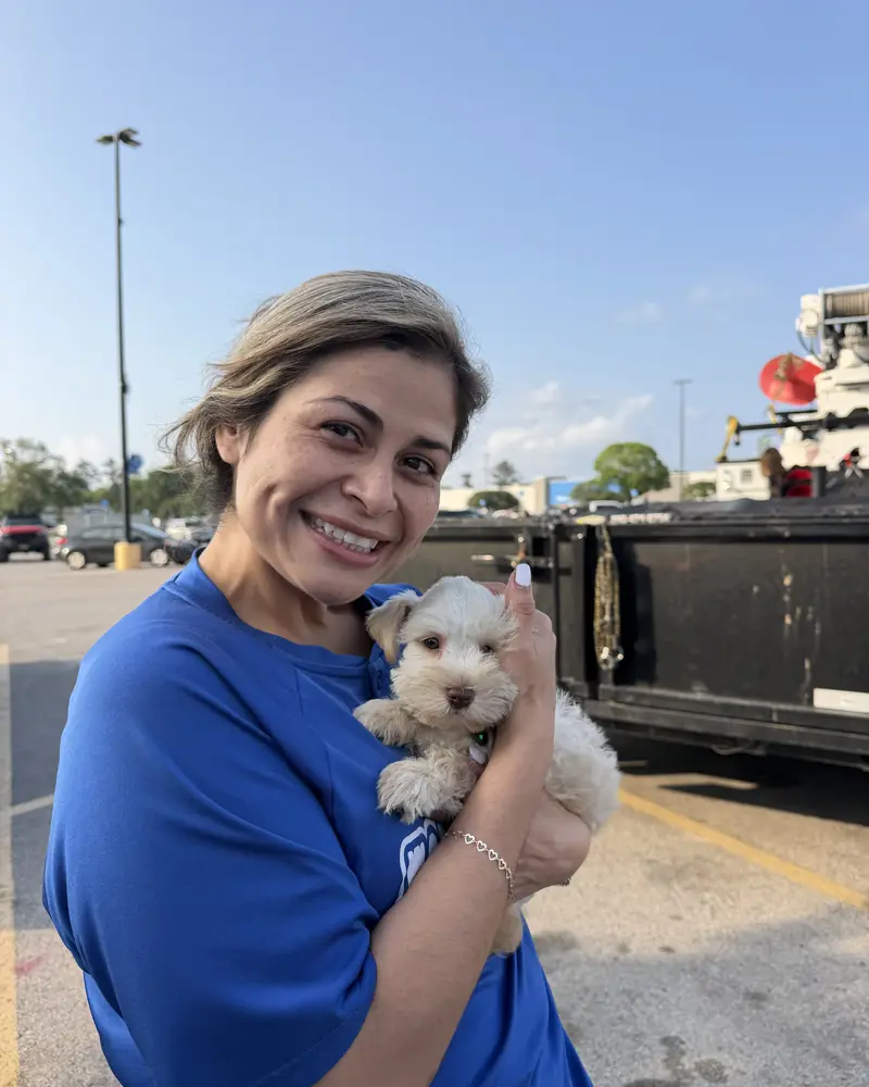 A woman in a blue shirt holding her new cream puppy against her shoulder in a car park.