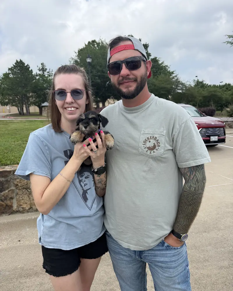 A couple standing in a car park, the woman holding their new salt-and-pepper puppy in both hands.