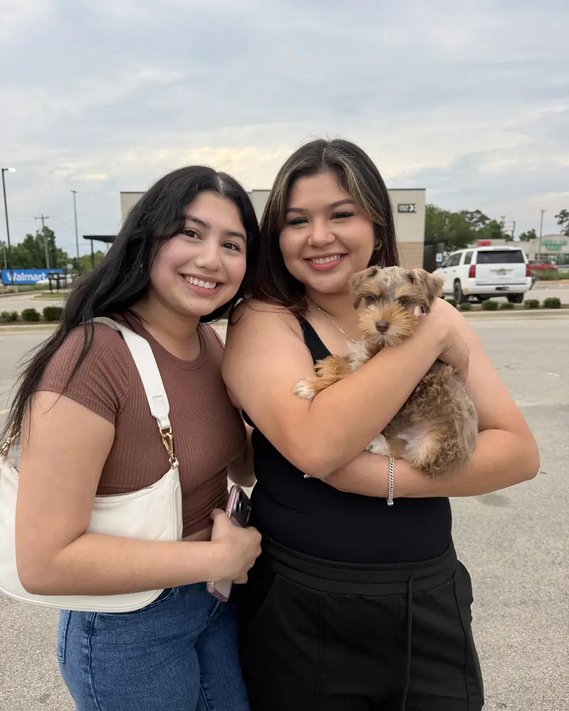 Two young women arm in arm in a car park, one holding their new cream puppy against her chest.