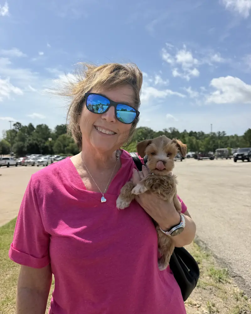 A woman in sunglasses and a pink shirt holding her new tan puppy up on her shoulder.