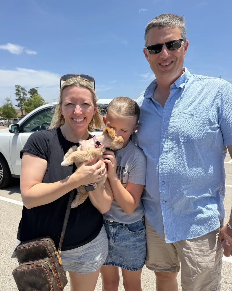 A couple and a young girl beside a white pickup, the girl leaning in to the new cream puppy in the woman&rsquo;s arms.