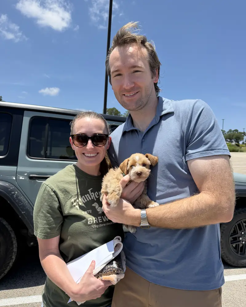 A couple in a car park, the man holding their new cream puppy while the woman carries the paperwork.