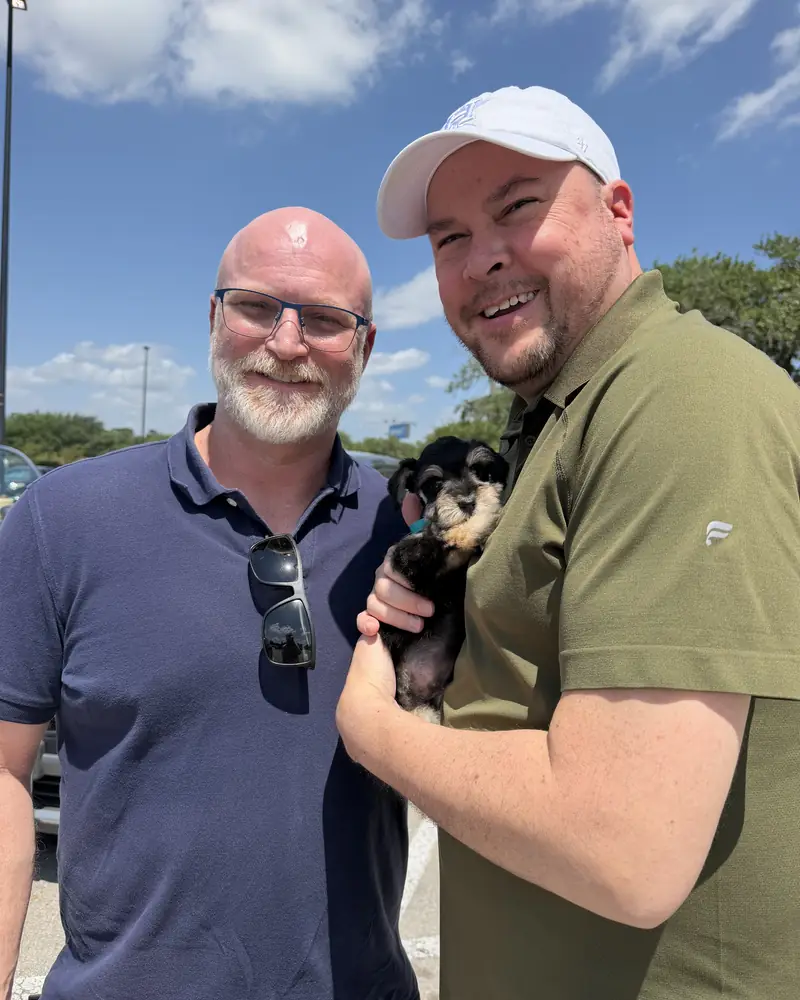 Two men in a sunny car park, one of them holding their new black-and-silver puppy against his chest.