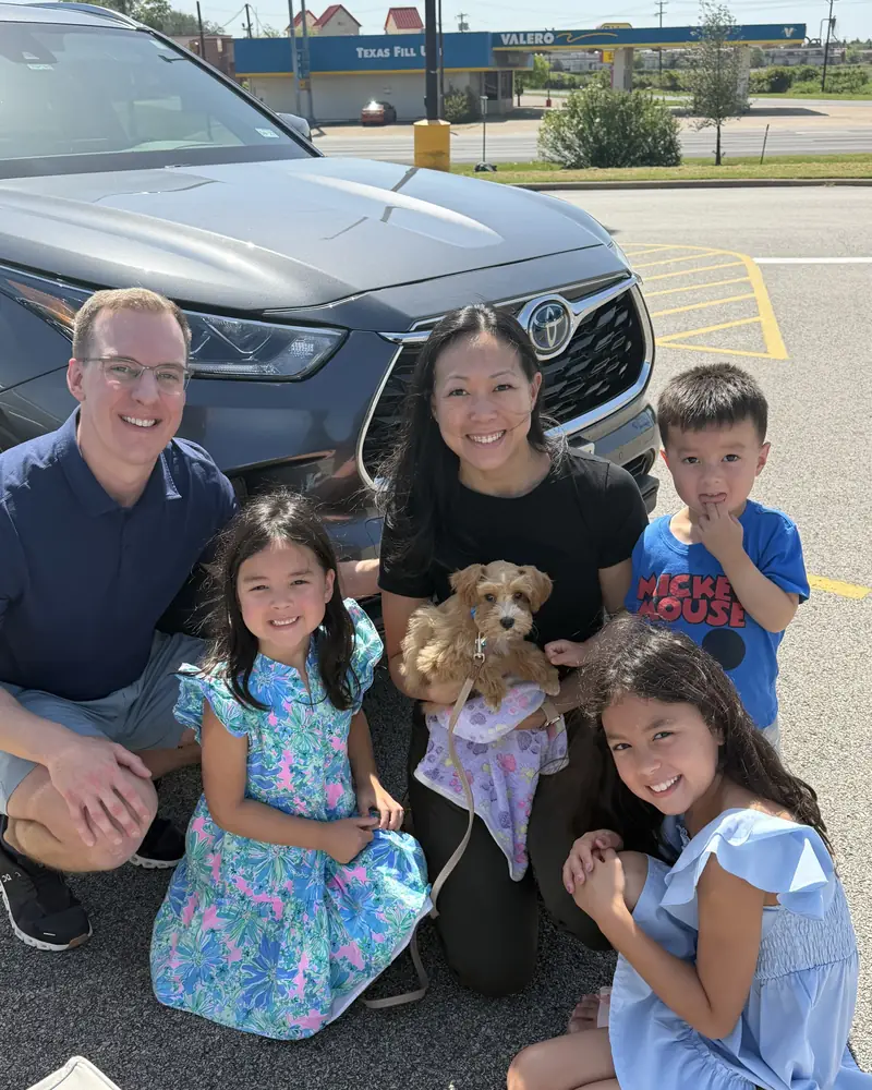 A family with three children kneeling in front of their car around their new tan puppy in a harness.