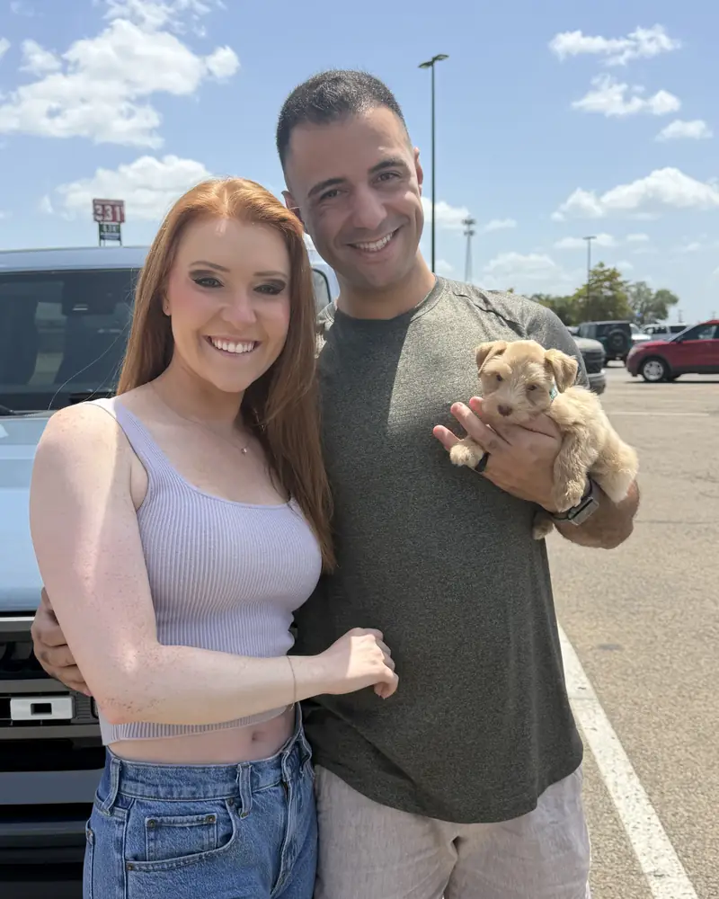 A couple standing by a pickup, the man holding their new cream puppy in one hand.