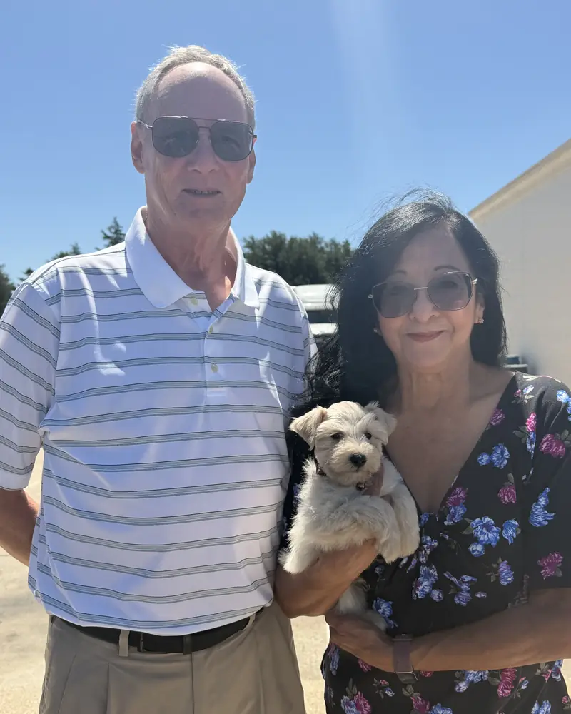 A couple outdoors in bright sun, the woman holding their new white puppy in both arms.