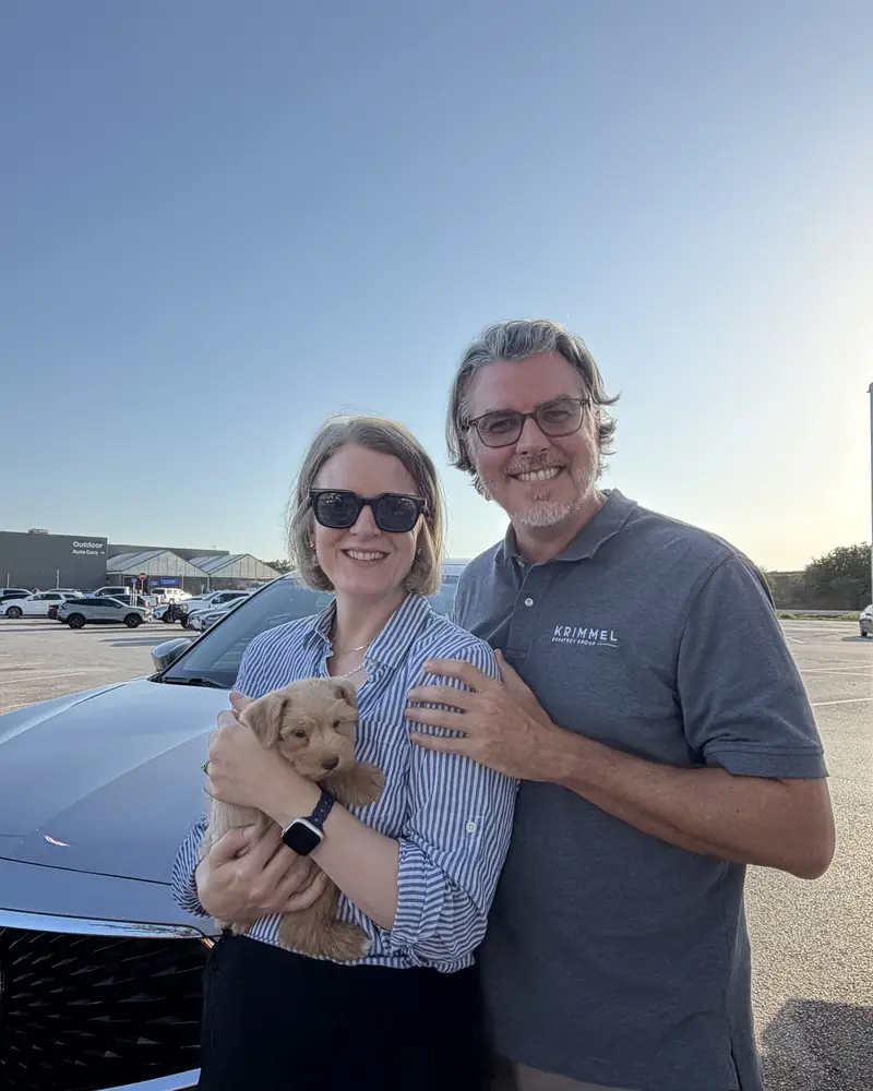 A couple beside a car at sunset, the woman holding their new cream puppy in both arms.