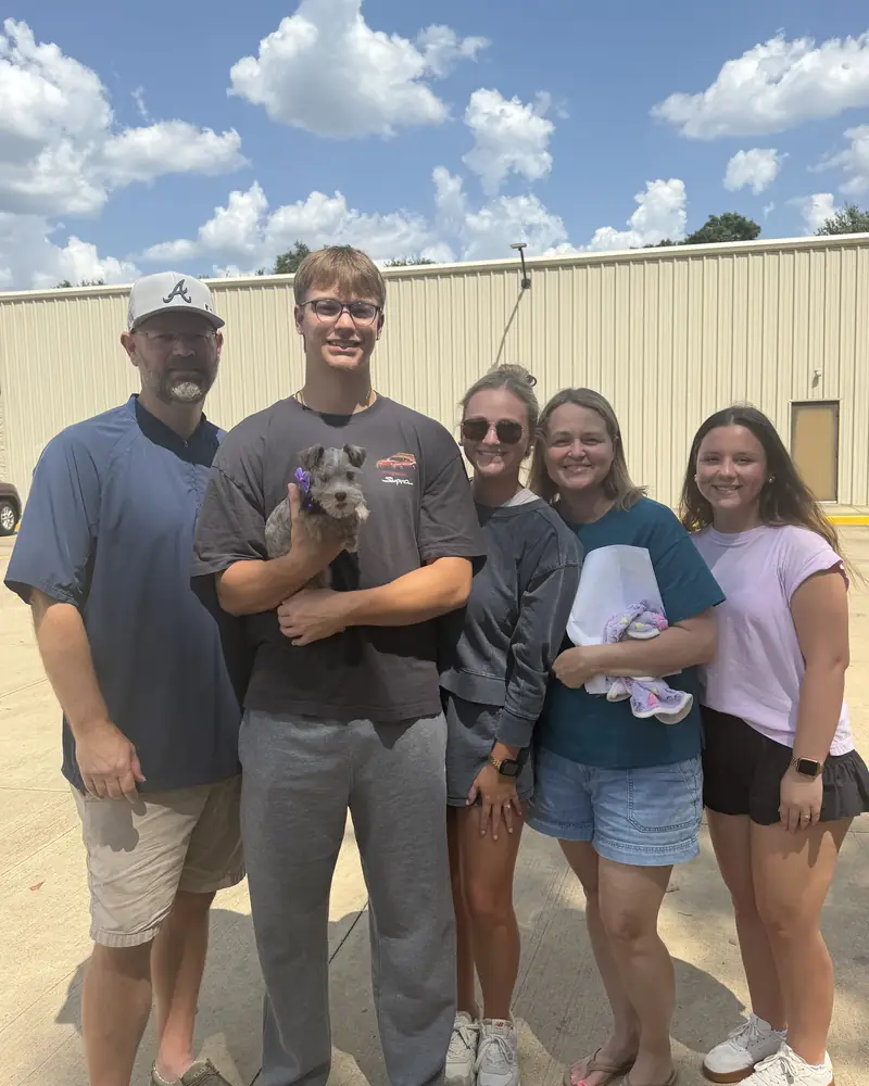 A family of six outside a metal building, one of them holding their new salt-and-pepper puppy in a purple bow.